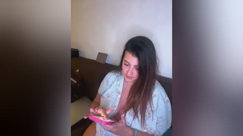 Media: Video of a young woman with long brown hair, wearing a light blue shirt, sitting on a couch eating pizza, surrounded by a dimly lit room with minimal decor.