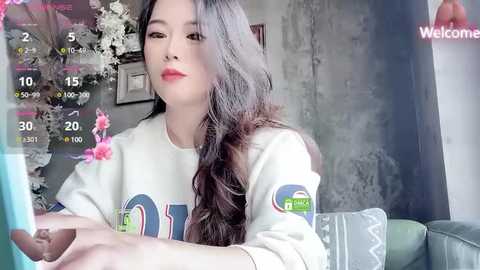 Media: Video of an Asian woman with long, wavy black hair, wearing a white sweater, sitting in a modern living room with gray walls, floral decor, and a digital calendar overlay.