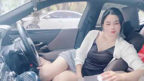 Media: Video of an Asian woman with fair skin, black hair, wearing a black V-neck top, white cardigan, and grey skirt, seated in a car, smiling, steering wheel in hand.