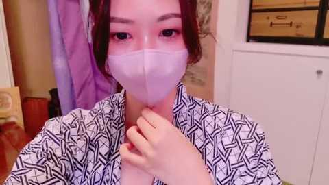 Media: Video of an East Asian woman with light skin, dark hair, and red lipstick, wearing a white mask, black patterned kimono, and holding her neck, in a cluttered room with wooden furniture.