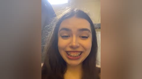 Media: A video of a young woman with straight brown hair and braces, smiling brightly, standing in front of a bathroom mirror with a tiled background.