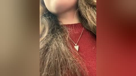 Media: Video of a woman's neck and shoulders, wearing a red sweater with a faux fur collar, and a gold heart necklace. Background is blurred in warm tones.