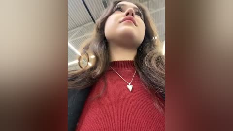 Media: A video of a young woman with long, wavy brown hair, wearing a red knit top and a heart-shaped necklace, looking up at a ceiling with a metal grid, taken from a low angle.