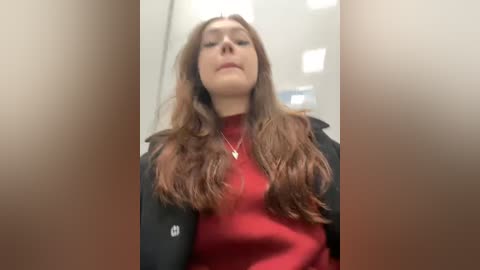 Media: A video of a young woman with long, wavy brown hair, wearing a red turtleneck sweater and black jacket, standing in an indoor setting with blurred, white-walled background.