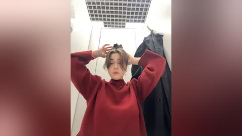 Media: Video of a young woman with light skin and short, tousled hair, wearing a red sweater, adjusting her hair in a changing room. Background features a grid ceiling and a black coat hanging nearby.