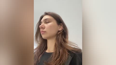 Media: Video of a young woman with long, dark brown hair, wearing a black top, standing in a plain, off-white room. She has a serene expression, eyes closed, and appears to be meditating.