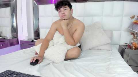 Media: Video of a young, shirtless Latino man with curly hair, sitting on a white bed, holding a pillow and remote control, in a modern, white-themed bedroom with purple accents.