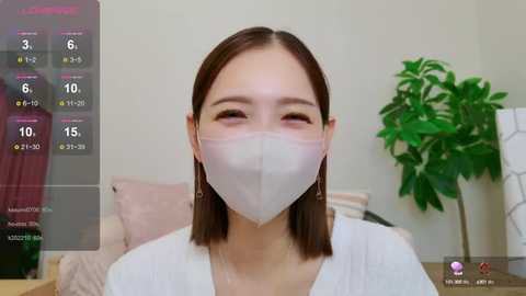 Media: Video of an Asian woman with shoulder-length brown hair, wearing a white mask and a white top, seated indoors against a plain background with a potted plant.