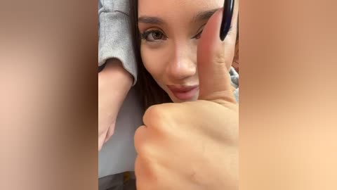 Media: A video of a young woman with light skin and brown eyes, wearing a gray top, giving a thumbs-up sign with her right hand, while the left arm is partially visible, suggesting a casual, confident pose.