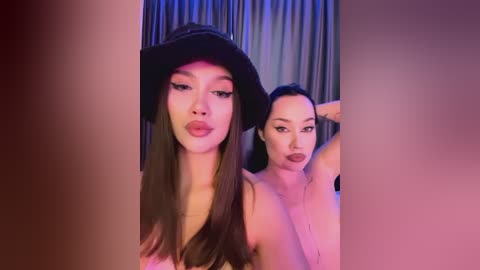 Media: Video of two women, one topless with a black hat, the other topless with dark hair, posing in a dimly lit room with curtains.