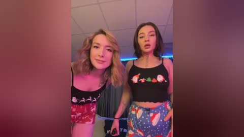 Media: Video of two young women with light skin, one with blonde hair and the other with dark hair, wearing festive tank tops and pajama pants. They stand in a dimly lit room with a tiled ceiling.