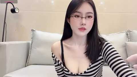 Media: Video of an East Asian woman with fair skin, straight black hair, and glasses, wearing a black-and-white striped top, sitting on a light gray couch in a modern living room.