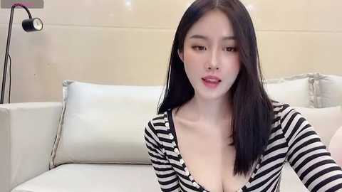 Media: Video of an East Asian woman with long black hair, fair skin, and wearing a black and white striped shirt, posing in a modern, minimalist living room with white furniture and a lamp.