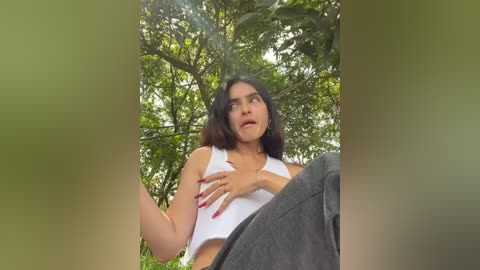 Media: Video of a woman with shoulder-length dark hair, wearing a white crop top and gray pants, standing outdoors in a lush, green forest. She has a surprised expression and is touching her chest with one hand.