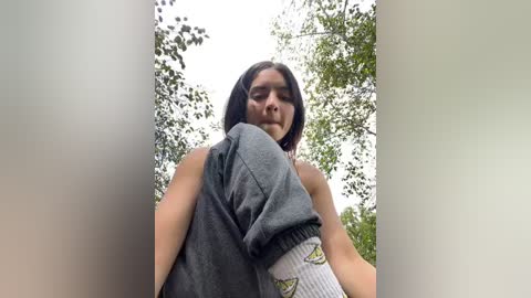 Media: Video of a young woman with medium skin tone, dark hair, and a light sleeveless top, sitting outdoors with her knees drawn up. Background shows green trees and a cloudy sky.