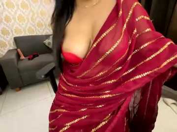 Media: Video of a woman in a red, gold-sequined sari with a red bra peeking through, standing in a modern living room with a gray couch and patterned wallpaper.