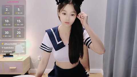 Media: A video of an Asian woman with long black hair in a Japanese schoolgirl cosplay outfit, adjusting her headband in a minimalist room with a calendar and wooden furniture.