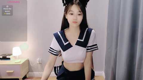 Media: Video of an East Asian woman with long black hair, wearing a revealing sailor-inspired costume with a cropped top and skirt. She sits on a bed with grey curtains in the background.