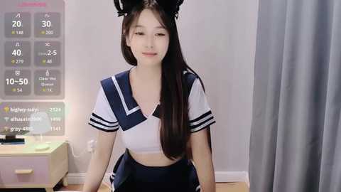 Media: Video of an East Asian woman in a Japanese schoolgirl cosplay outfit, wearing black cat ears, sitting on a bed with a digital display in the background.