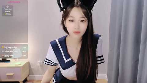 Media: A video of a young Asian woman with long black hair, wearing a schoolgirl outfit, black cat ears, and a blue and white sailor collar. She stands indoors near a window, with a TV and white walls in the background.