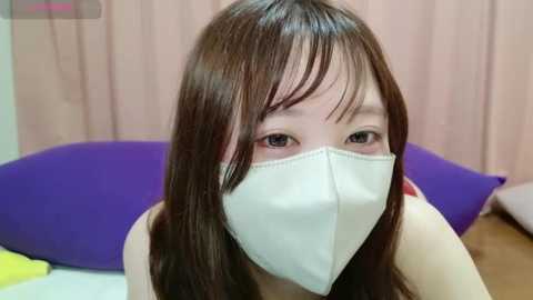 Media: A close-up video of an Asian woman with long brown hair, wearing a white surgical mask, sitting indoors on a bed with purple and yellow pillows.