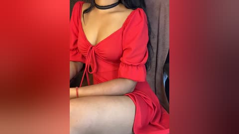 Media: Video of a woman in a red dress with a deep V-neckline and puffed sleeves, sitting on a chair. She wears a black choker and has long black hair. The background is a gradient of reds, enhancing the vibrant dress.