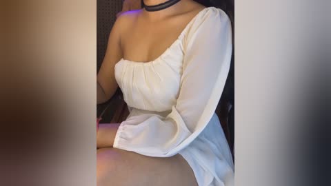 Media: Video of a woman with medium skin tone wearing a white, sheer, off-shoulder top and black choker, seated on a black chair in a dimly lit room.