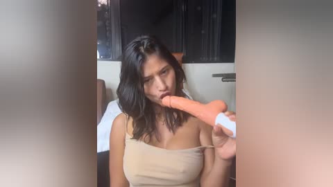 Media: Video of an Asian woman with shoulder-length black hair, wearing a beige tank top, giving a blowjob to a realistic, flesh-colored dildo. The setting appears to be a dimly-lit room with dark curtains.