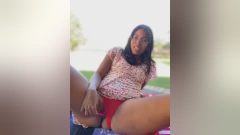 Media: Video of a young South Asian woman with long black hair, wearing a floral top and red shorts, sitting outdoors with legs spread, revealing her vulva.