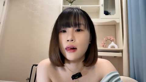 Media: Video of an Asian woman with straight, shoulder-length black hair and pale skin, wearing a light off-shoulder top and a black necklace. She's in a beige-walled room with a white shelf holding decorative items.