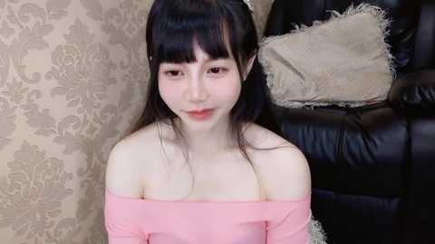 Media: A video of an Asian woman with long black hair, wearing a pink off-shoulder top, sitting on a black leather couch. The background features beige damask wallpaper and a beige pillow.