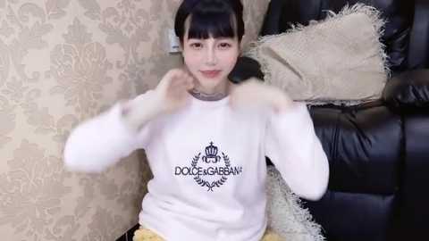 Media: Video of a smiling East Asian woman with straight black hair, wearing a white \"Dolce & Gabbana\" sweatshirt, sitting on a black leather couch with beige pillows.