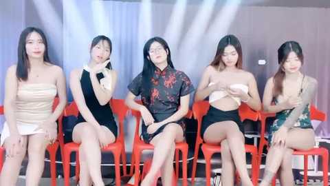 Media: Video of five Asian women, seated in a row, on red plastic chairs, wearing various revealing outfits, in a brightly lit room with white curtains.