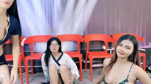 Media: Video of three Asian women in casual attire; two sit, one stands, against a backdrop of red plastic chairs and a white curtain.