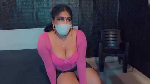 Media: Video of a woman with dark hair, wearing a pink long-sleeve top, black shorts, and a blue face mask, sitting on a bed in a dimly lit room with a dark wall and a black chair.