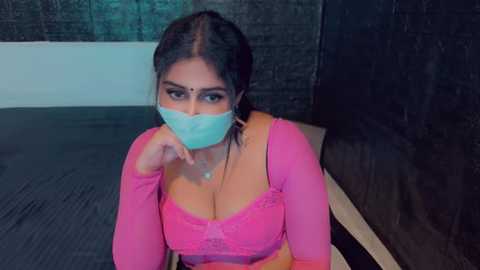 Media: Video of a young woman with light brown skin, dark hair, and large breasts, wearing a pink lace bra and mask, sitting on a dark couch in a dimly lit room.