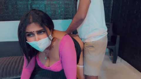 Media: Video of a South Asian woman in a pink top, face mask, and black shorts, being penetrated from behind by a man in a white tank top and beige shorts in a dimly-lit room.