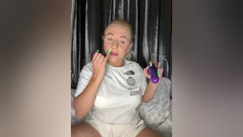 Media: Video of a blonde woman with fair skin, wearing a white t-shirt and shorts, sitting on a bed with patterned sheets. She holds a green vibrator and a purple dildo, smiling, in a casual, intimate setting.