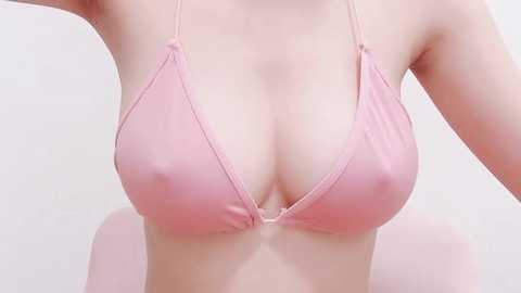 Media: Video of a woman wearing a light pink, string bikini top with thin straps, revealing her cleavage. She has fair skin and is seated against a plain white background.