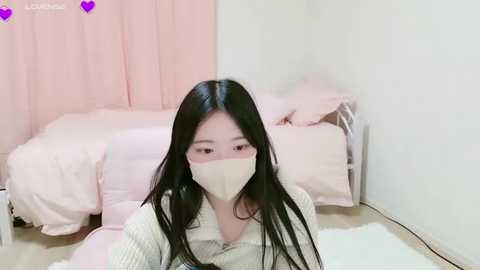 Media: Video of an Asian woman with long black hair, wearing a beige mask, white sweater, and pink pajamas, sitting on a white rug in a pastel-pink bedroom.