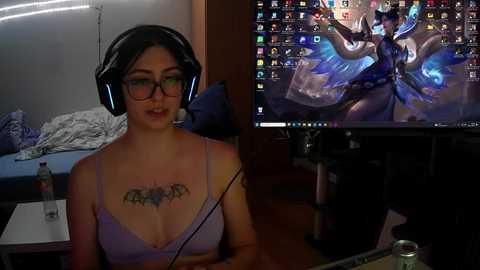 Media: Video of a young woman with glasses, black hair, wearing a lavender bra, tattoo on chest, playing video game with large headset. Background shows a cluttered room with bed and computer.