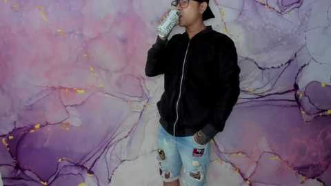 Media: Video of a young man with light brown skin, wearing a black zip-up hoodie, light blue jeans with patches, and a backward baseball cap. He drinks from a water bottle in front of a pastel abstract mural with pink, purple, and white tones.