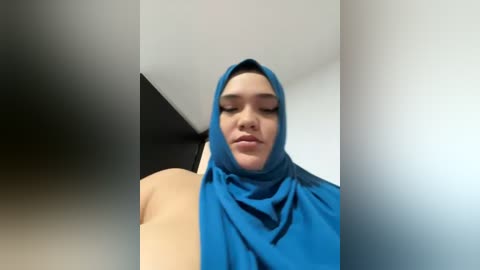 Media: Video of a woman with light skin, wearing a blue hijab, standing against a white wall. She has a neutral expression and is captured from a low angle, emphasizing the hijab's texture and her facial features.