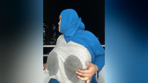 Media: Video of a large, round woman with a blue hijab, seated outdoors at night. She has a gray sweater with large circular designs. The background shows city lights in the distance.