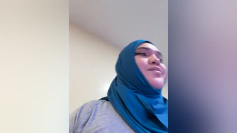 Media: Video of a young woman with fair skin, wearing a teal hijab and light grey top, standing against a plain white wall. She has a neutral expression and is leaning against a column.