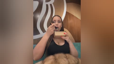 Media: Video of a curvy, light-skinned woman with long brown hair, wearing a black top, eating a slice of cake with a fork, in a room with wooden paneling and a circular white design on the wall.