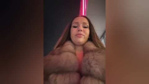 Media: Video of a woman with light skin and long brown hair, wearing a pink fur coat and a pink hat, looking down. The background is dimly lit.