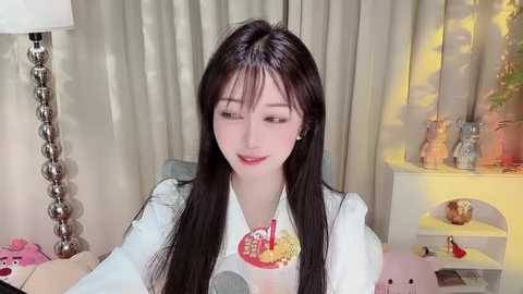 Media: A video of a young Asian woman with long black hair, fair skin, and delicate features, wearing a white shirt with a graphic design. She sits in a cozy room with a lamp, curtains, and decorative items.