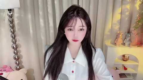 Media: Video of a young East Asian woman with long black hair, fair skin, and red lipstick, wearing a white blouse, seated in a cozy, well-lit room with beige curtains and a lamp.