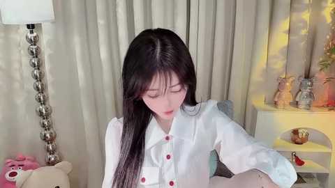 Media: Video of a fair-skinned East Asian woman with long black hair, wearing a white blouse, seated indoors. Background features beige curtains, a lamp, stuffed toys, and a shelf with decorative items.